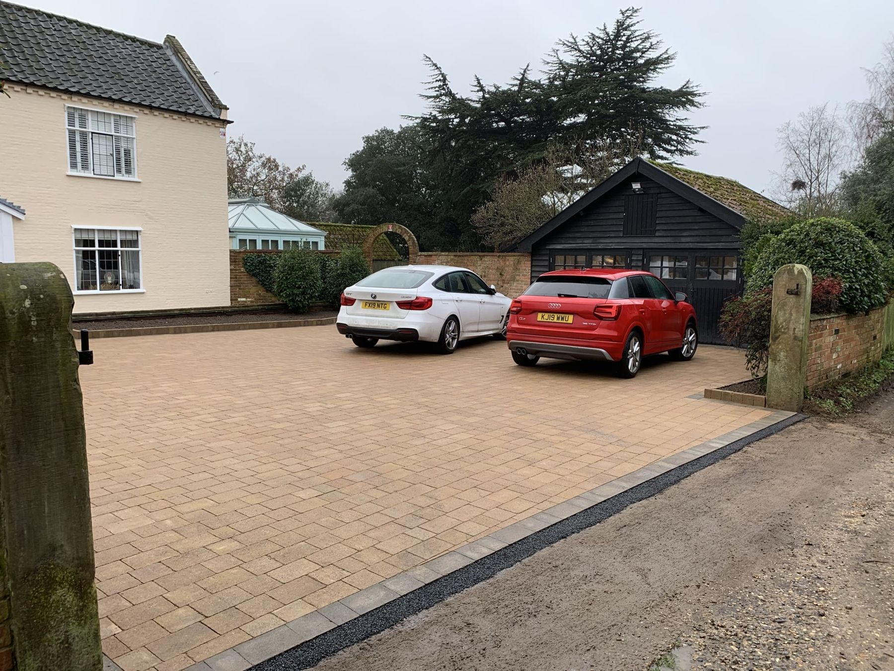 Block paved driveway with two cars in front of a cream house and garage