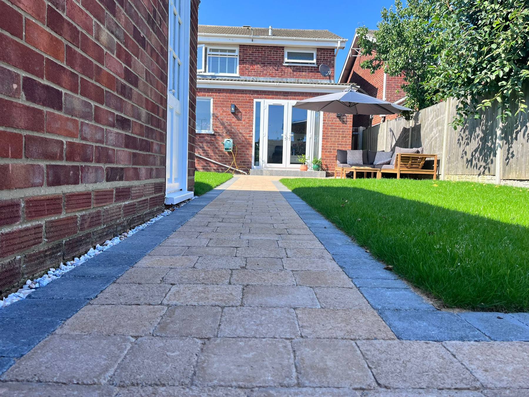 Block paving path running beside a new lawn to a patio with a parasol behind a red brick house