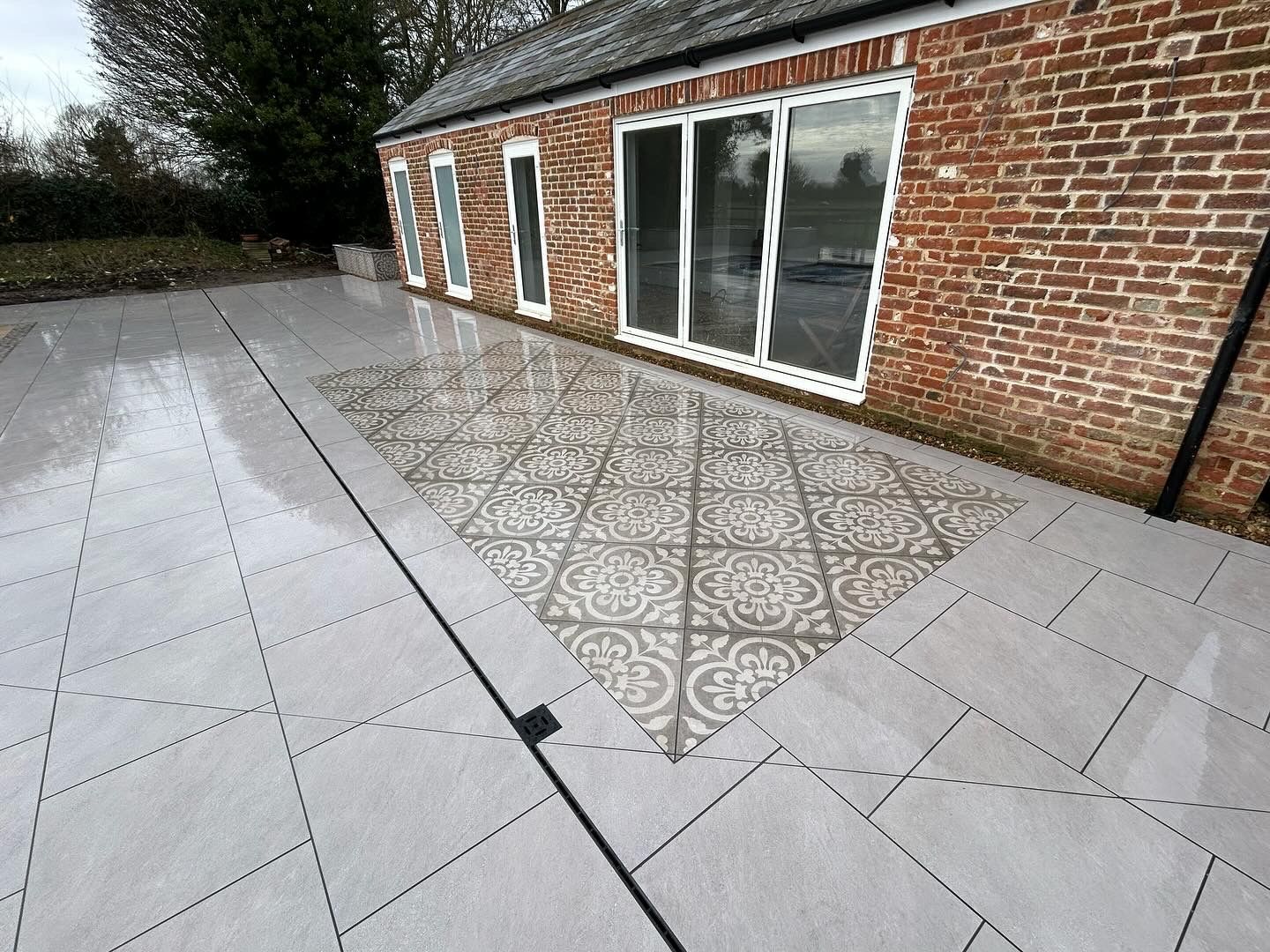 Large grey porcelain patio with a patterned tile inlay beside a brick building
